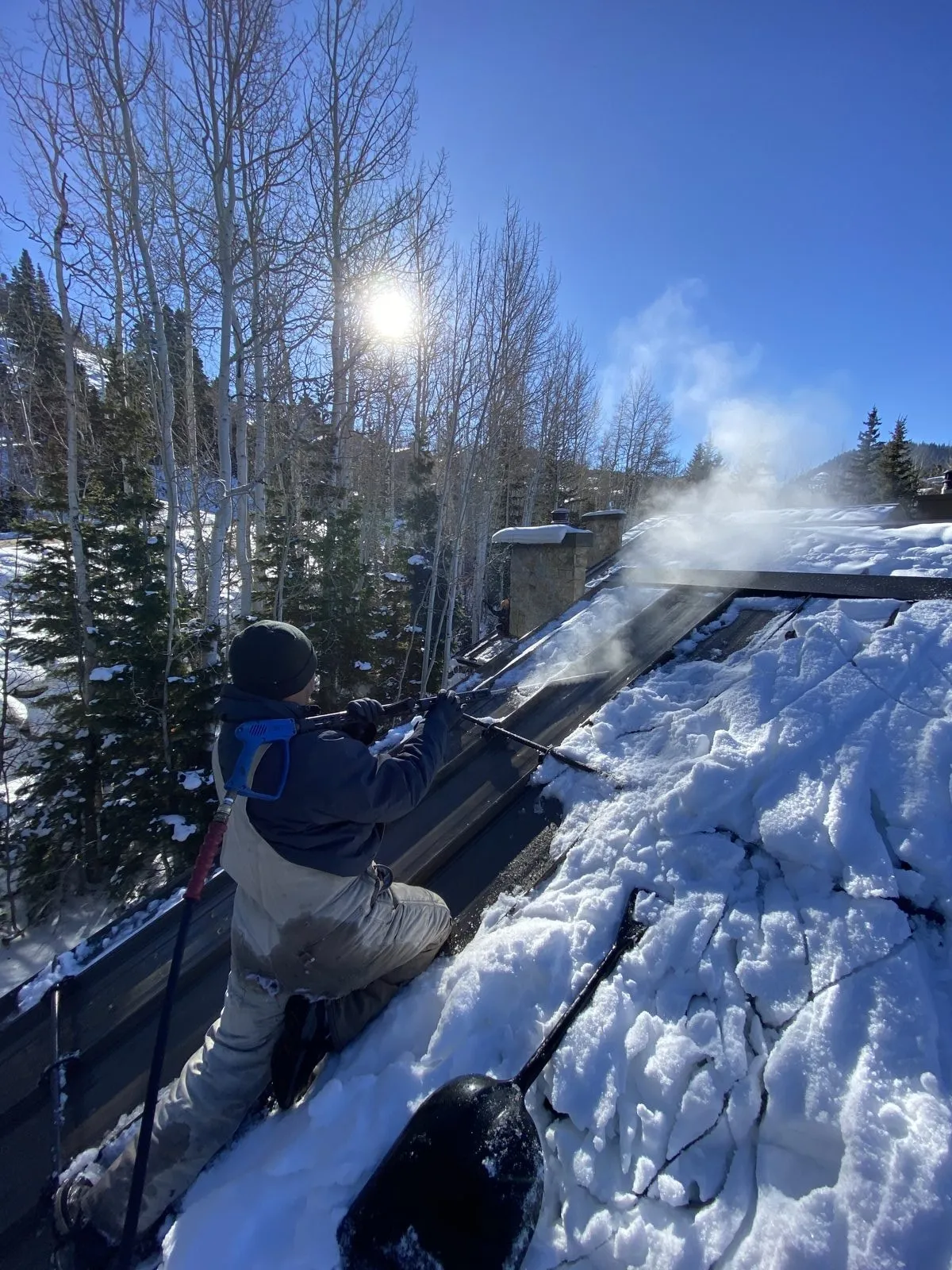 Alaska Heat Cable crew member steaming an ice dam off a metal roof in bright winter sun